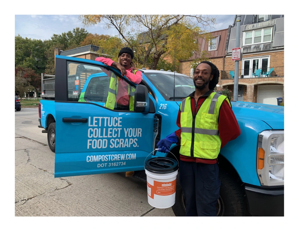 Two City workers for the Curbside Composting Pilot Program stand near their sky blue pickup truck and smile while their picture is taken.